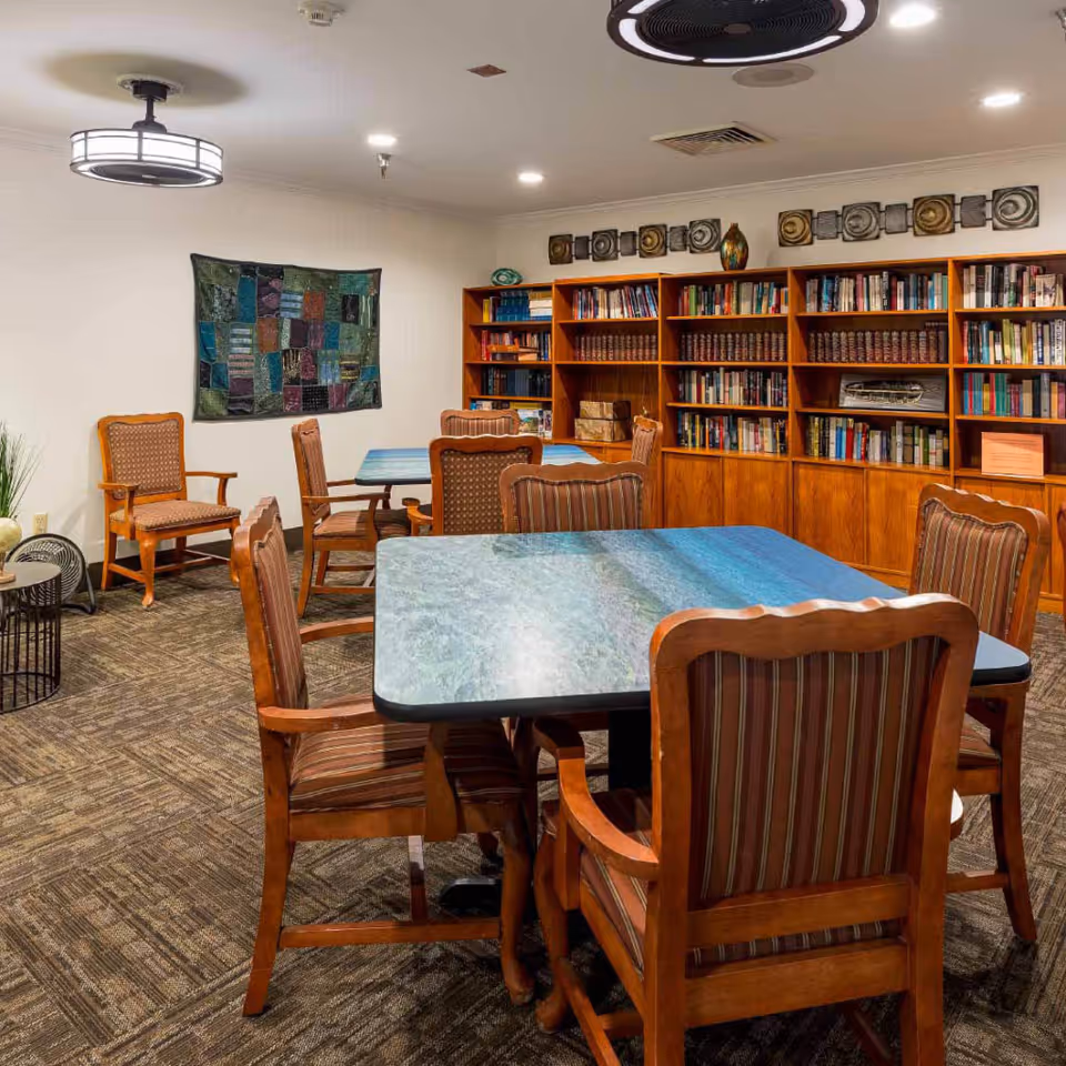 A cozy reading or activity room with wooden tables and cushioned chairs arranged around them. There is a large wooden bookshelf filled with books along the back wall, a decorative wall hanging, and modern ceiling lights illuminating the space.