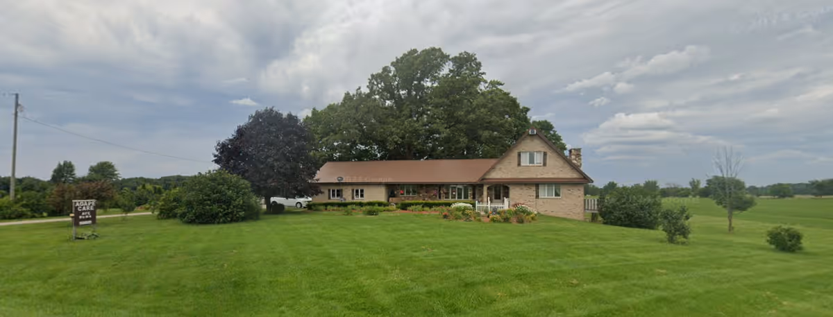 A single-story brick building with a brown roof surrounded by a large, well-maintained grassy lawn and several trees under a cloudy sky. A sign on the left side of the image reads 'Agape Care'.