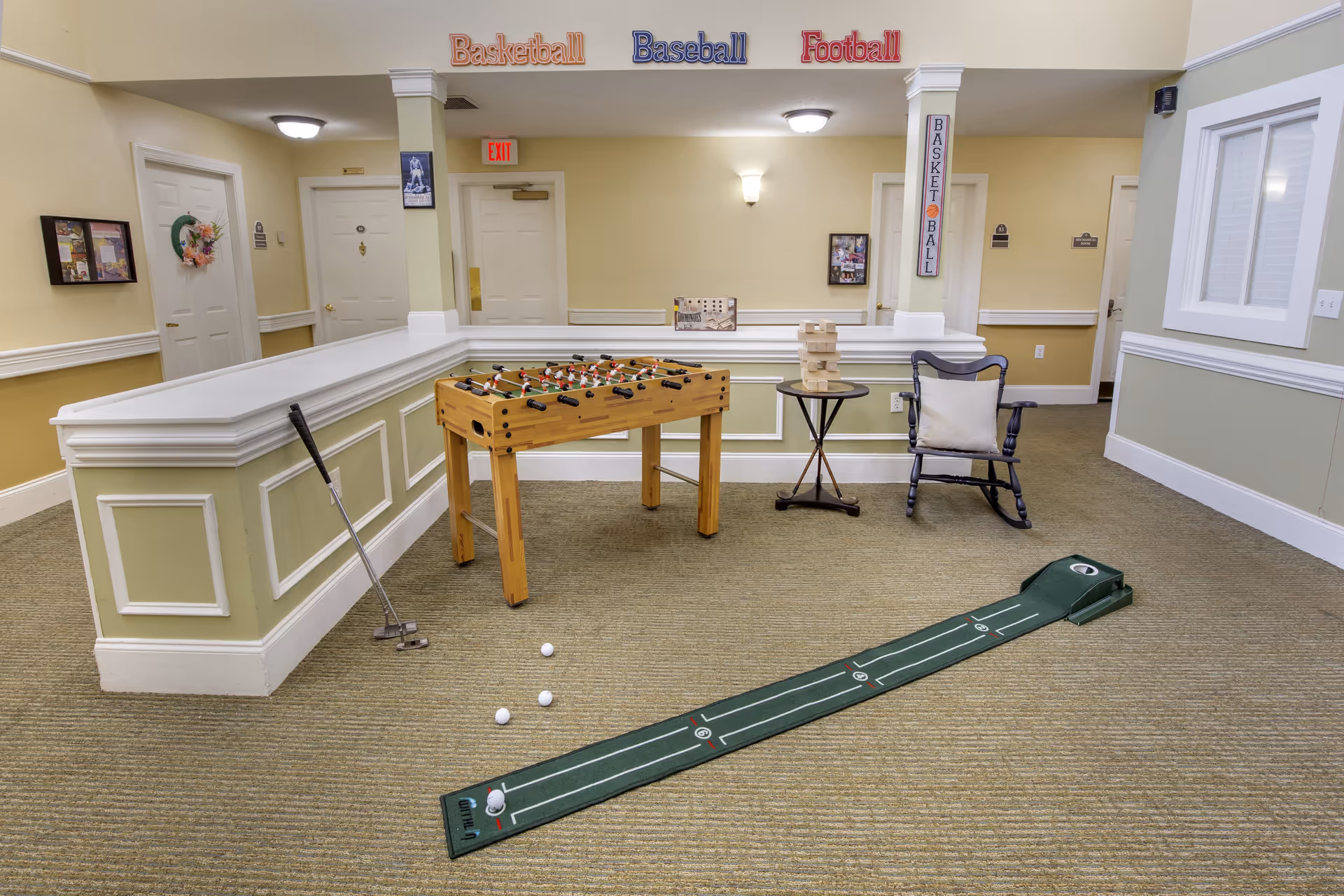 A recreational room with a foosball table, a small putting green with golf balls and a putter, a rocking chair with a cushion, and a small round table with a Jenga game on it. The walls are light green and beige with white trim, and there are sports-themed decorations on the walls including signs for Basketball, Baseball, and Football.