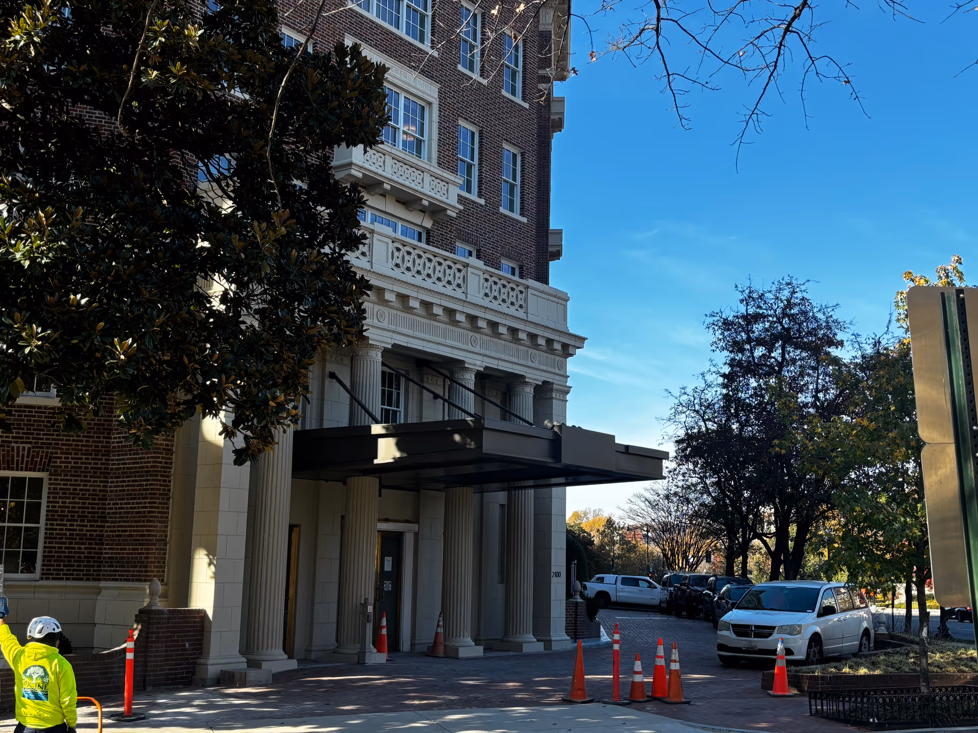 Entrance of a multi-story brick building with classical architectural details including columns and decorative stonework. Several orange traffic cones are placed near the entrance and along the driveway. A person wearing a bright yellow jacket and white helmet is visible on the left side. Trees and parked cars line the street under a clear blue sky.