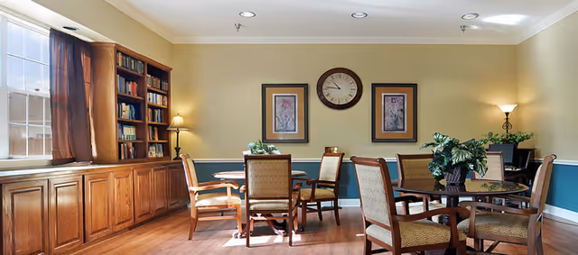 A well-lit common room with wooden flooring, several round tables surrounded by cushioned chairs, a large wooden bookshelf filled with books, two framed pictures and a clock on the wall, and two lamps providing warm lighting.