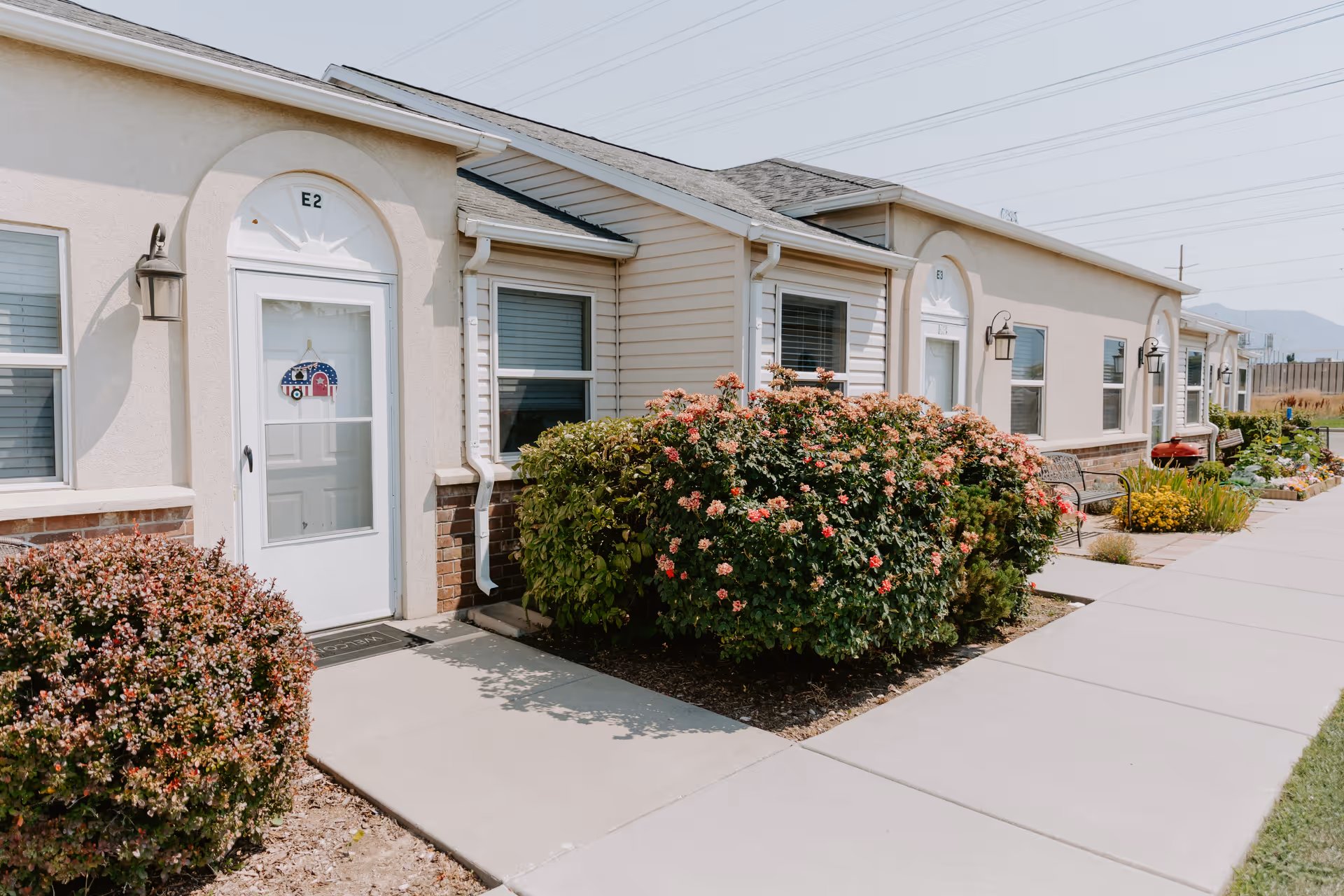 Exterior view of a single-story retirement community building with white doors labeled E2 and E3, windows with blinds, and well-maintained bushes and flowers along a concrete walkway under a clear sky.