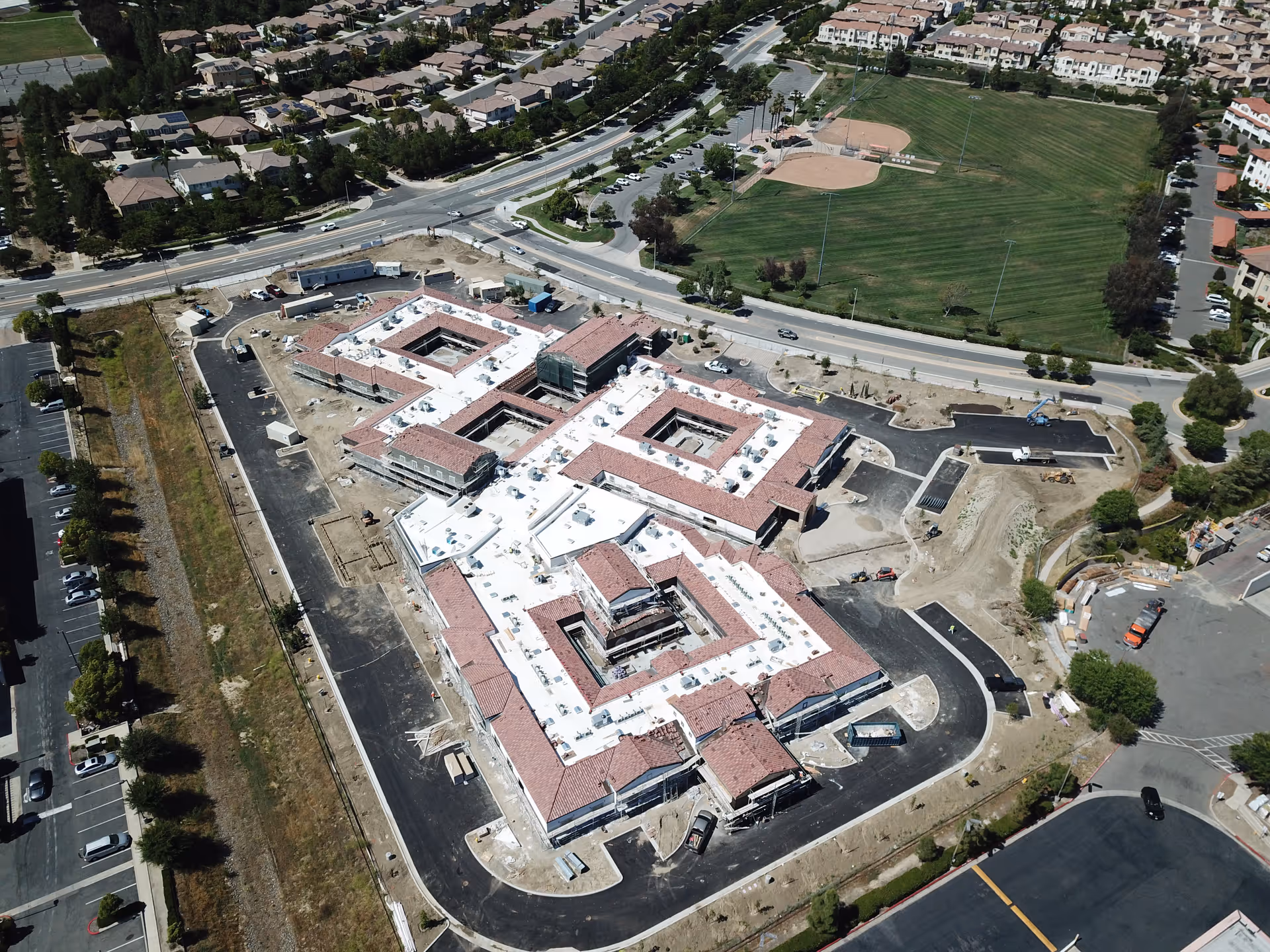 Aerial view of a large building under construction surrounded by paved roads and parking areas, with residential neighborhoods and a baseball field visible in the background.