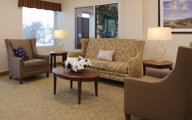 A cozy seating area in a senior living facility with a patterned beige sofa, two brown armchairs, a wooden oval coffee table with white flowers, two round side tables each with a lamp, a window with blinds, and a framed landscape painting on the wall.