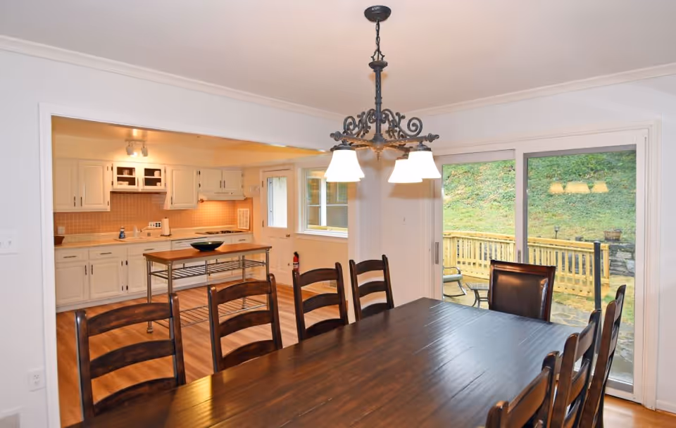 Dining area with a dark wooden table and eight chairs under a decorative chandelier light fixture. In the background, there is a kitchen with white cabinets, a small island with a bowl on top, and a sliding glass door leading to an outdoor patio with a wooden railing and green grass beyond.
