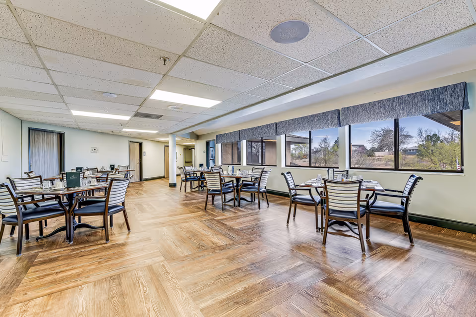 Bright communal dining room with multiple set round tables and chairs beside large windows overlooking trees.