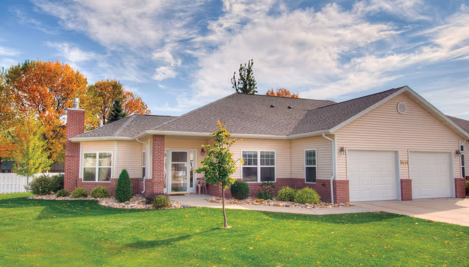 Single-story residential-style building with an attached two-car garage, front porch, landscaped lawn, and autumn trees.