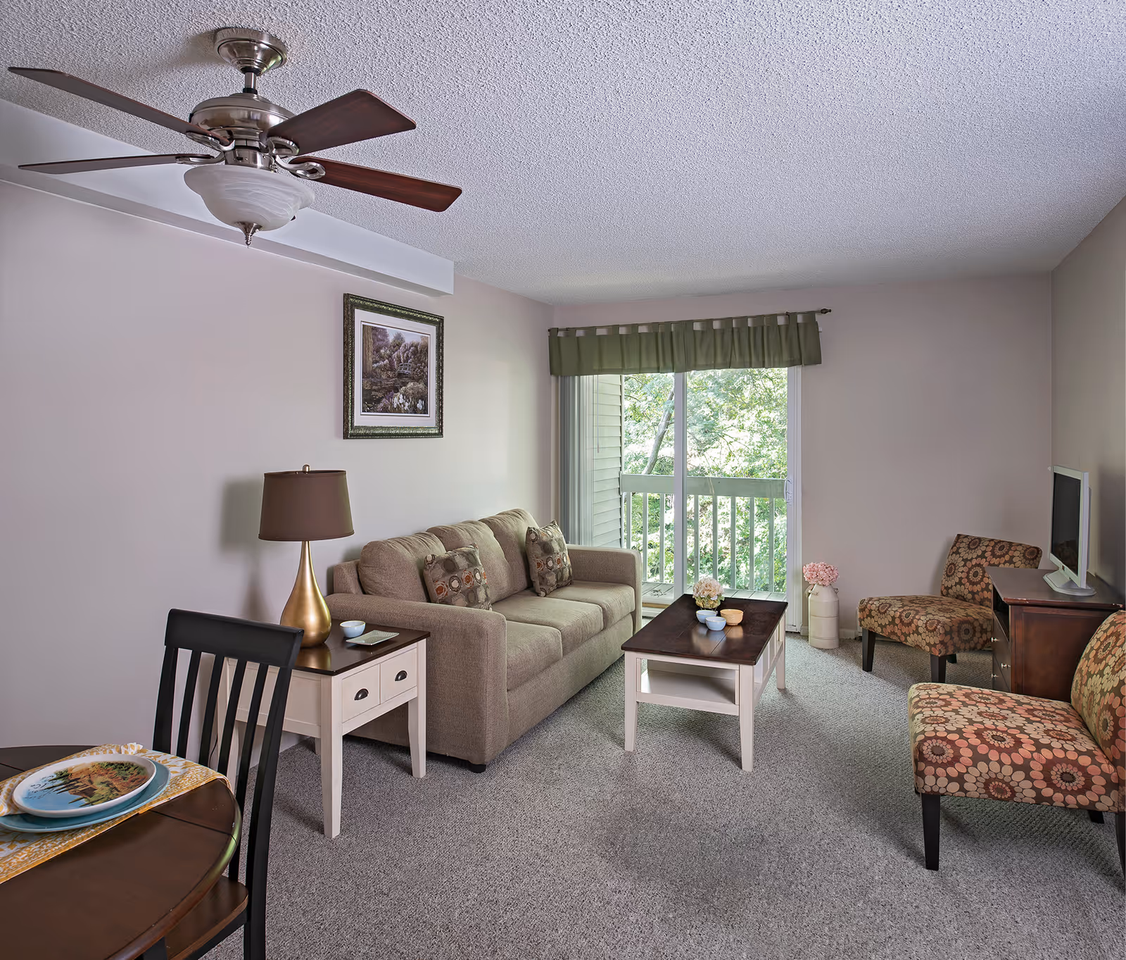 A cozy living room with a beige sofa adorned with patterned cushions, a wooden coffee table with decorative items, two patterned armchairs, a small TV on a wooden stand, a side table with a lamp, a ceiling fan, and a sliding glass door leading to a balcony with greenery outside.