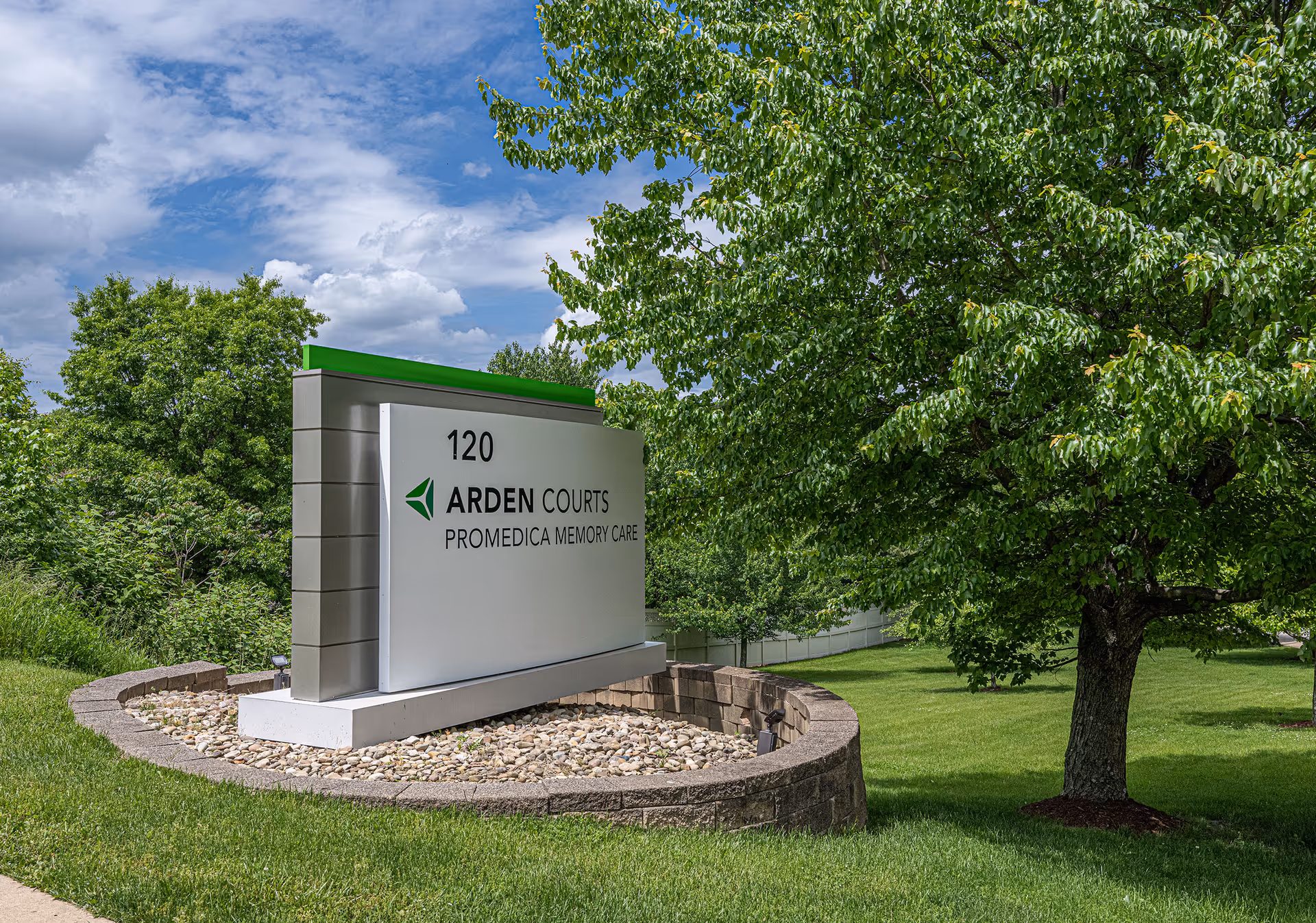 Outdoor view of a sign for Arden Courts ProMedica Memory Care Community at 120, surrounded by green grass, trees, and a partly cloudy sky.
