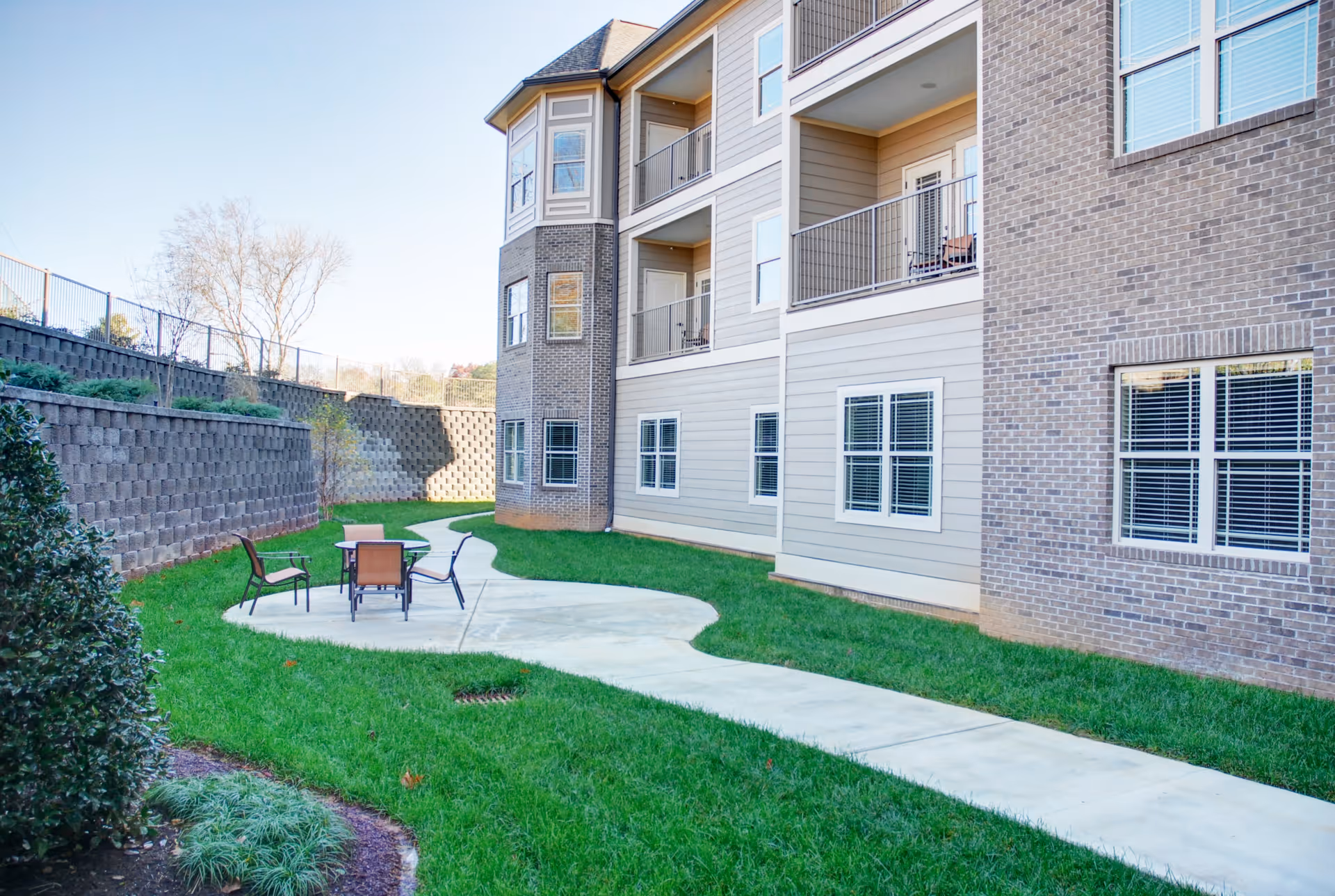 Outdoor area of a senior living facility with a concrete pathway leading to a small patio set with four chairs on a grassy lawn. The building has multiple floors with balconies and windows, and there is a retaining wall with plants on the left side.