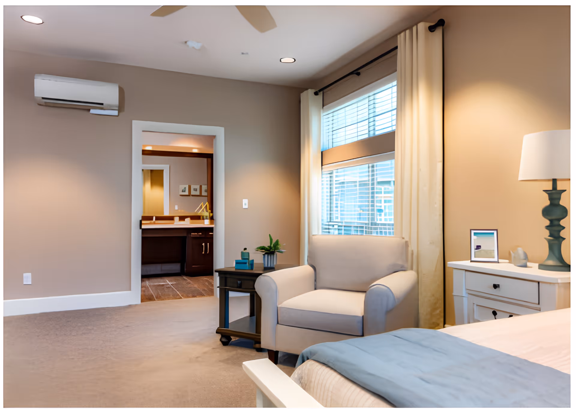A cozy bedroom with a beige armchair next to a window with white curtains and blinds. There is a nightstand with a lamp, a small framed picture, and decorative items. The bed has a blue blanket at the foot. In the background, an open doorway reveals a bathroom with a vanity and mirror.