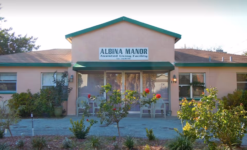 Front exterior view of Albina Manor Assisted Living Facility, a single-story building with a green roof trim, a small porch with chairs, and a garden with bushes and flowers in front.