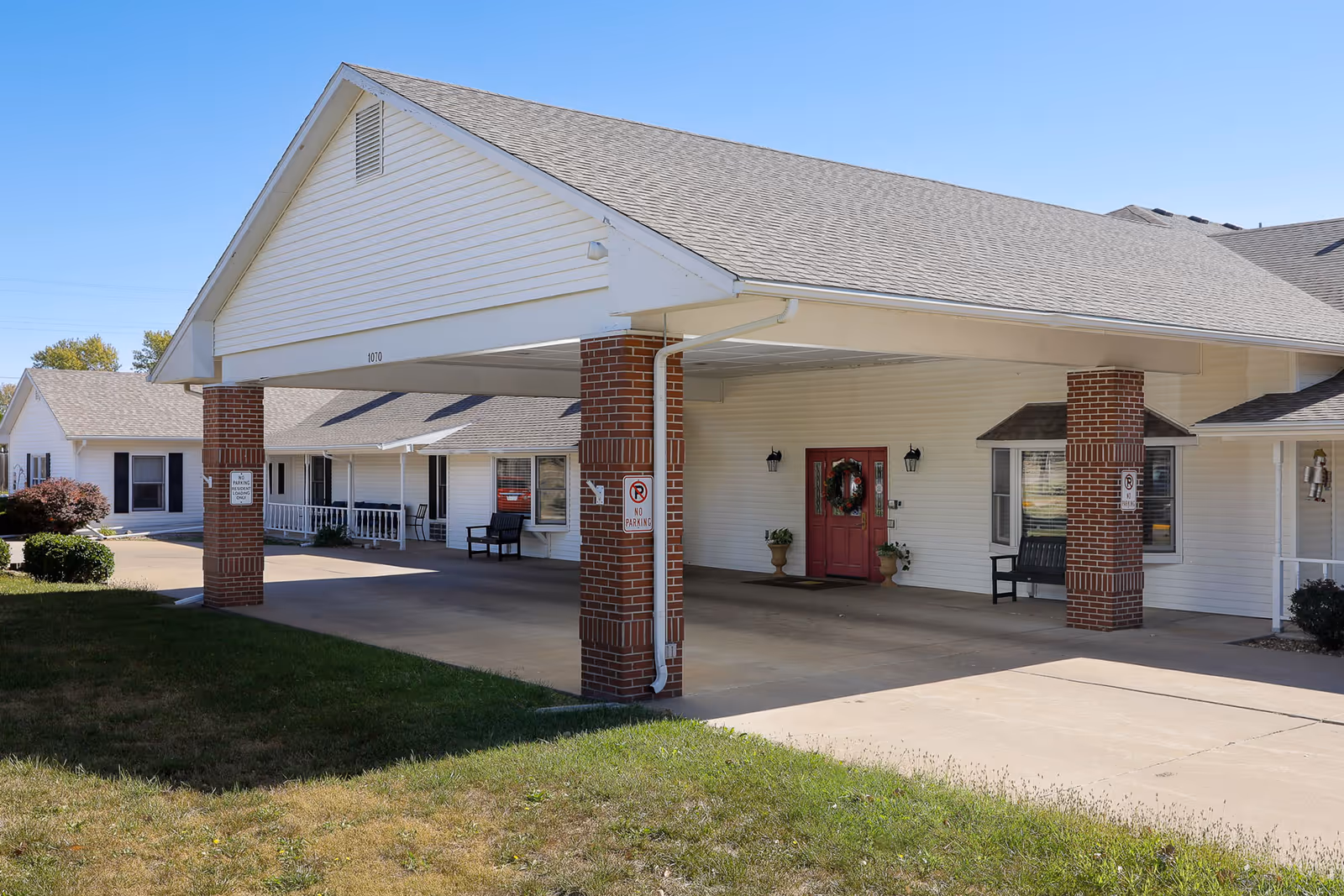 Covered front entrance with brick pillars, a drive-through porte-cochère, and red double doors on a single-story assisted living building.