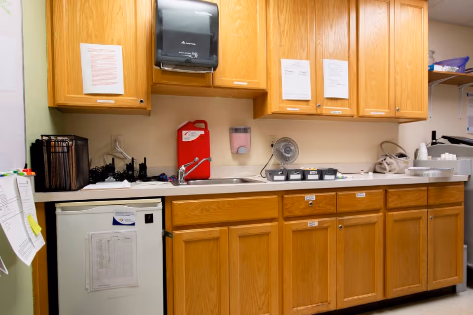 A small kitchenette area with wooden cabinets above and below a countertop. On the countertop are a small sink, a red sharps disposal container, a small fan, and various small containers. A paper towel dispenser is mounted on the wall above the sink. There is a mini refrigerator below the counter with papers attached to its door. Several papers and notes are taped to the cabinet doors and walls.