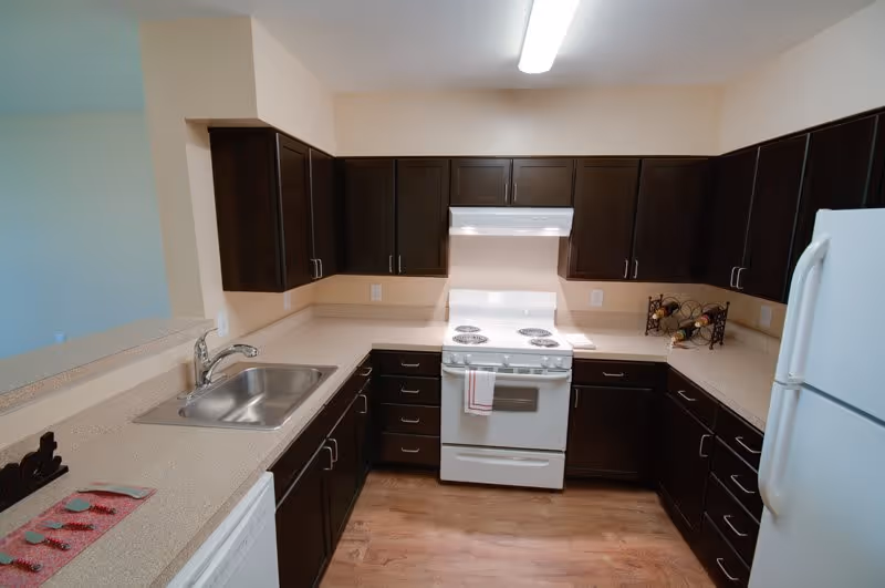 A modern kitchen with dark brown cabinets, beige countertops, a white electric stove with an oven, a white refrigerator, a stainless steel sink, and a wooden floor. There is a wine rack on the countertop and a dish towel hanging on the oven handle.