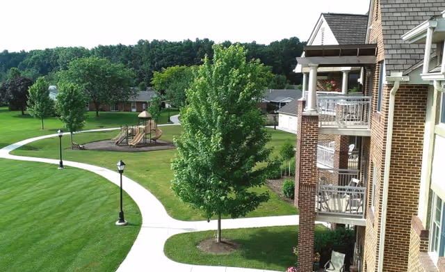 View of a senior living facility's outdoor area featuring a well-maintained lawn, a paved walking path, several trees, a playground with slides, and a brick building with balconies on the right side.