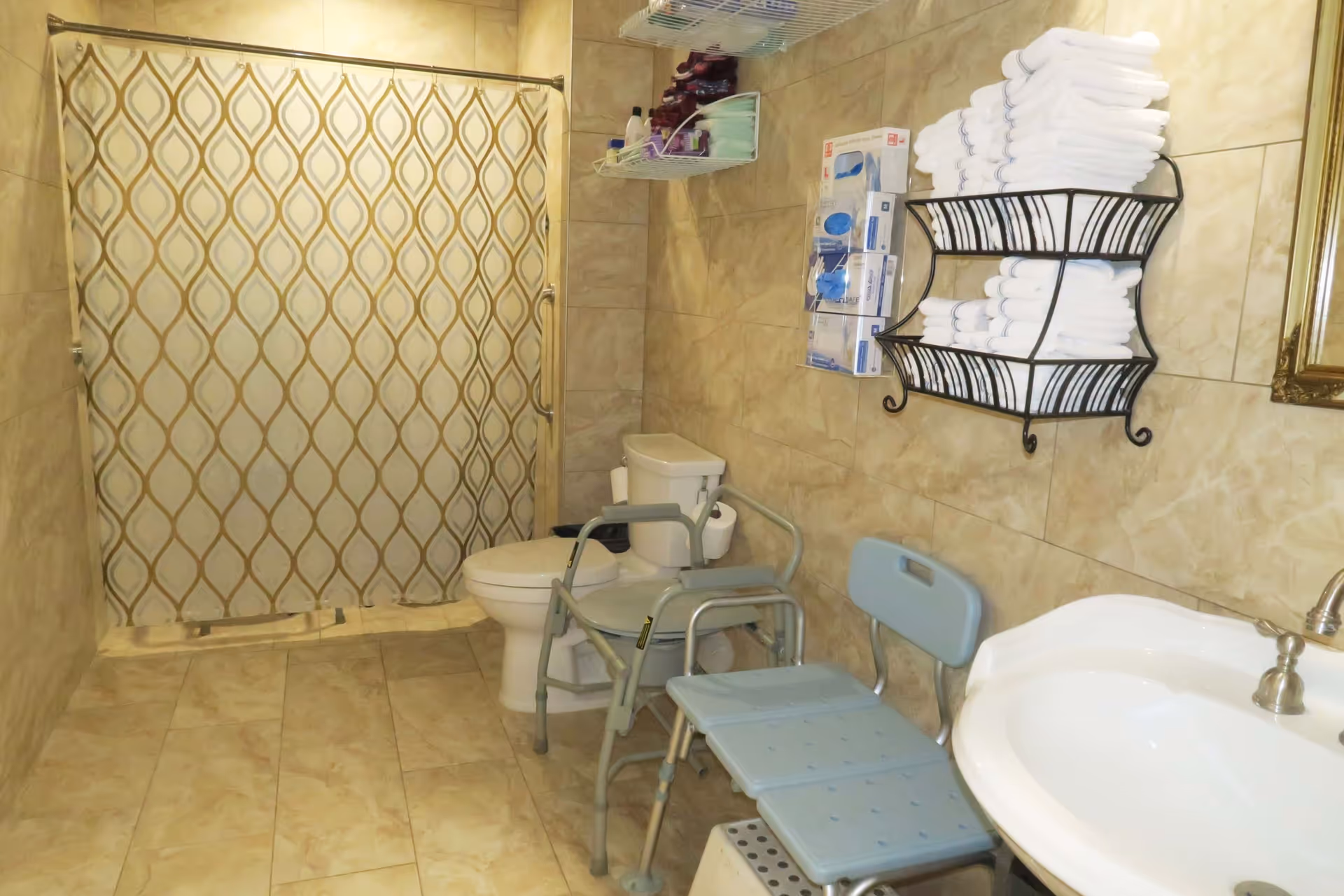 Bathroom with beige tiled walls and floor, a white toilet, a shower area with a patterned curtain, a white sink with a silver faucet, a metal rack holding neatly folded white towels, and a blue shower chair with a backrest and armrests.