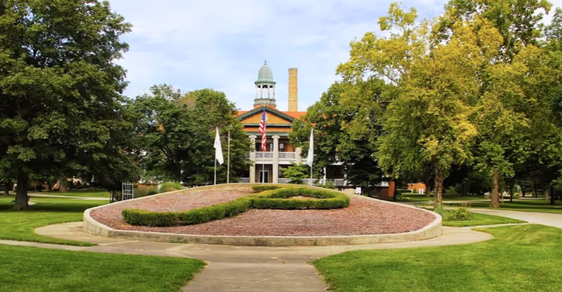A large historic building with a cupola and columns, partially obscured by trees, with a landscaped circular garden featuring trimmed bushes and an American flag in front, surrounded by green lawns and paved walkways under a partly cloudy sky.