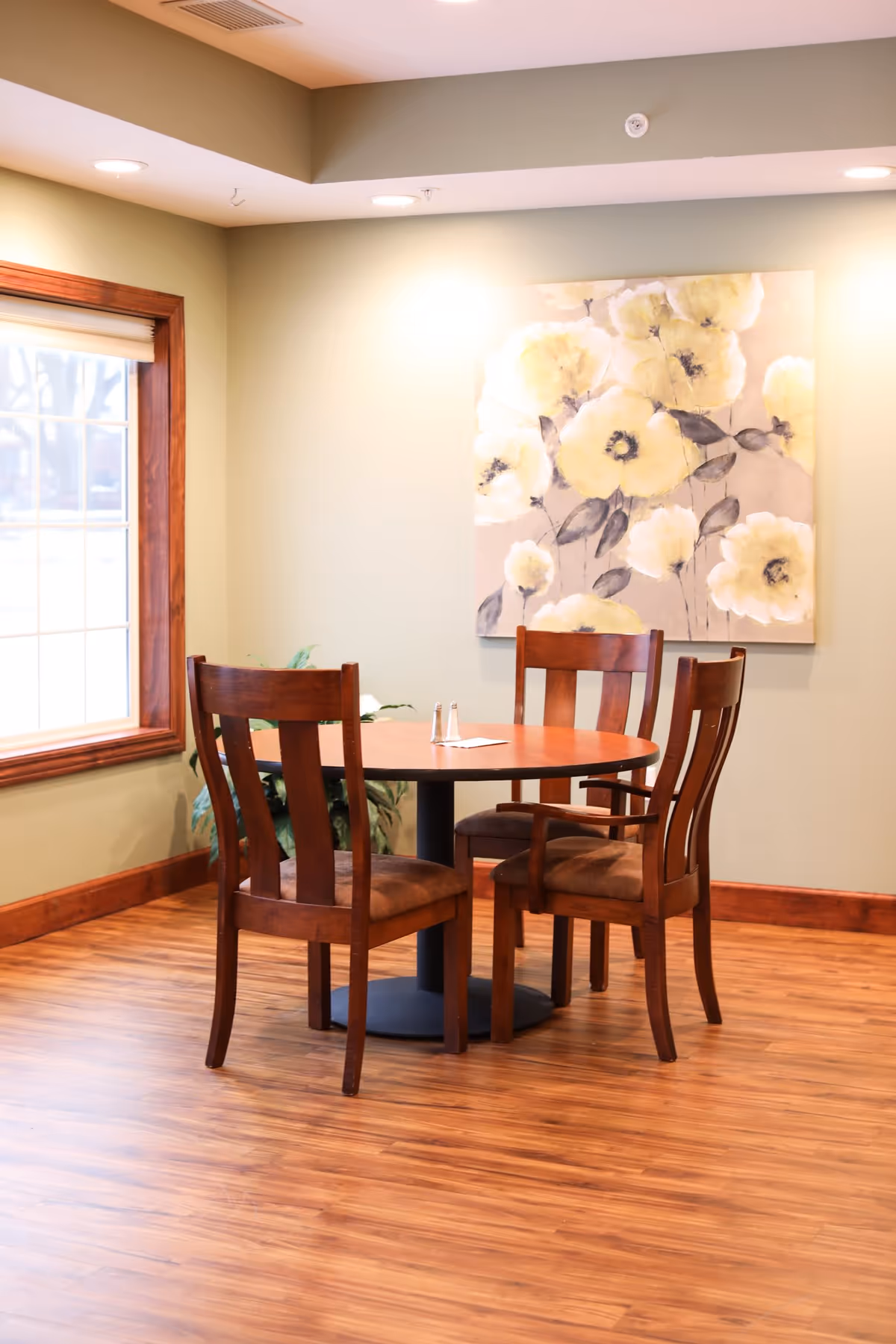 A small round wooden table with four wooden chairs arranged around it in a room with light green walls and wood flooring. A large painting of yellow flowers hangs on the wall behind the table, and a window with wooden trim is visible to the left.
