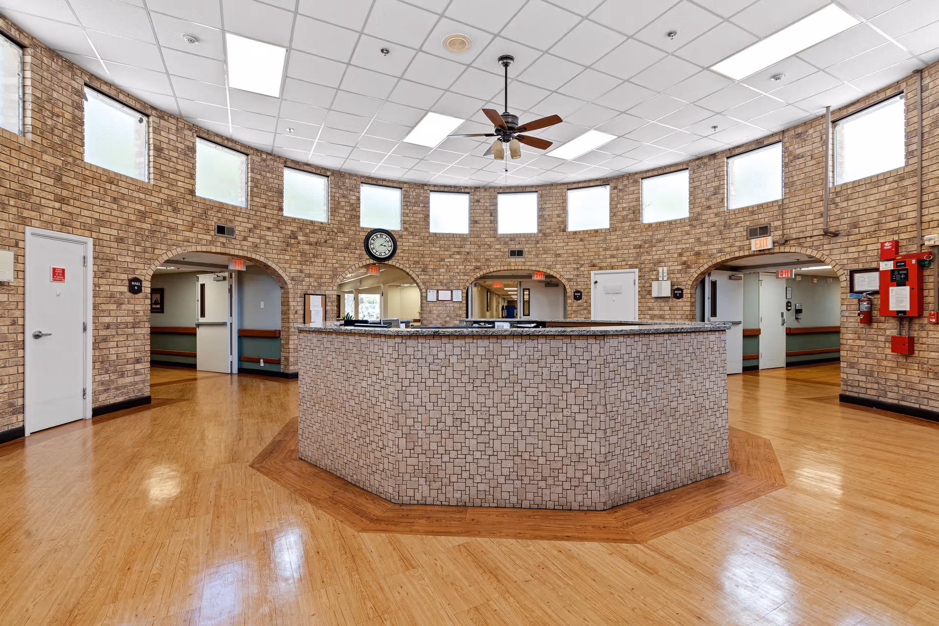 Interior view of a senior living facility reception area with a large octagonal front desk in the center. The walls are made of light brown brick with several frosted windows near the ceiling. There are three arched doorways behind the desk leading to hallways. The floor is wooden, and a ceiling fan with lights hangs above the desk. Various safety and fire equipment are mounted on the walls.