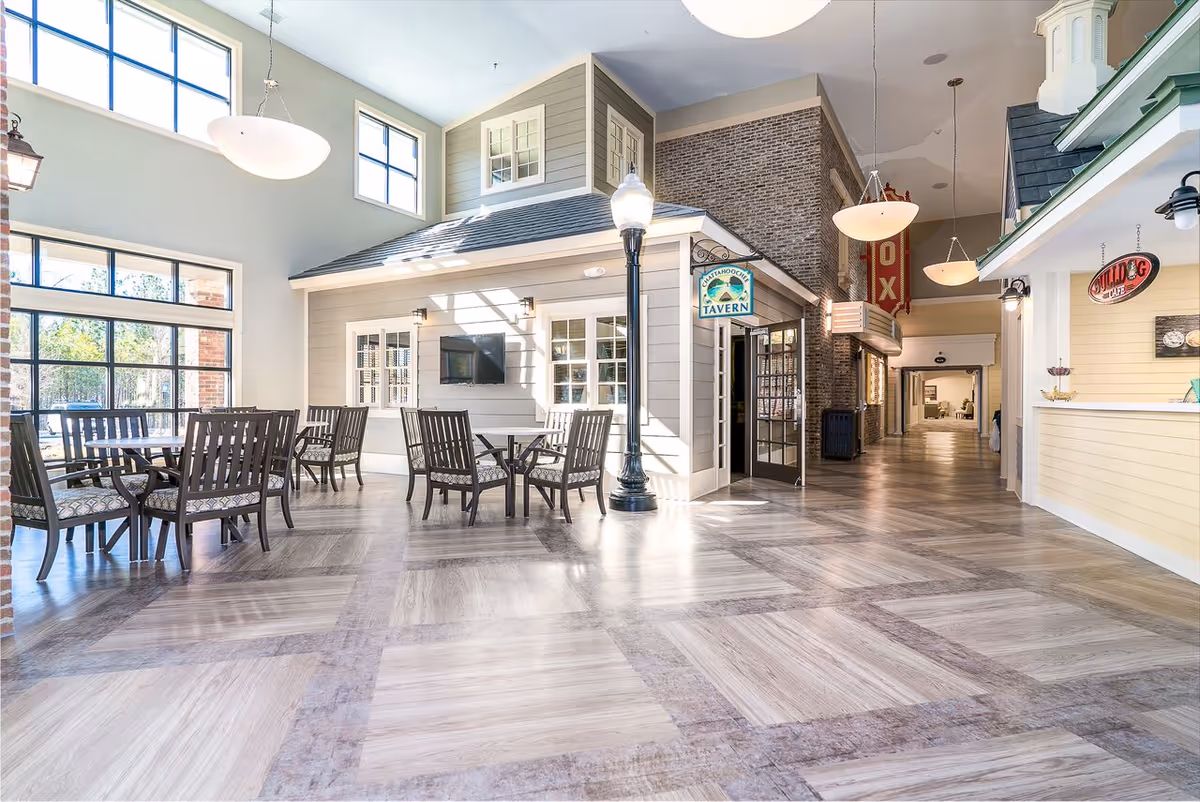 Bright and spacious interior common area with high ceilings and large windows letting in natural light. Several tables and chairs are arranged for seating. The space features a small building-like structure labeled 'Chattahoochee Tavern' with a TV mounted on the wall. There are hanging light fixtures and signs for '10X' and 'Bulldog Cafe' visible in the hallway.