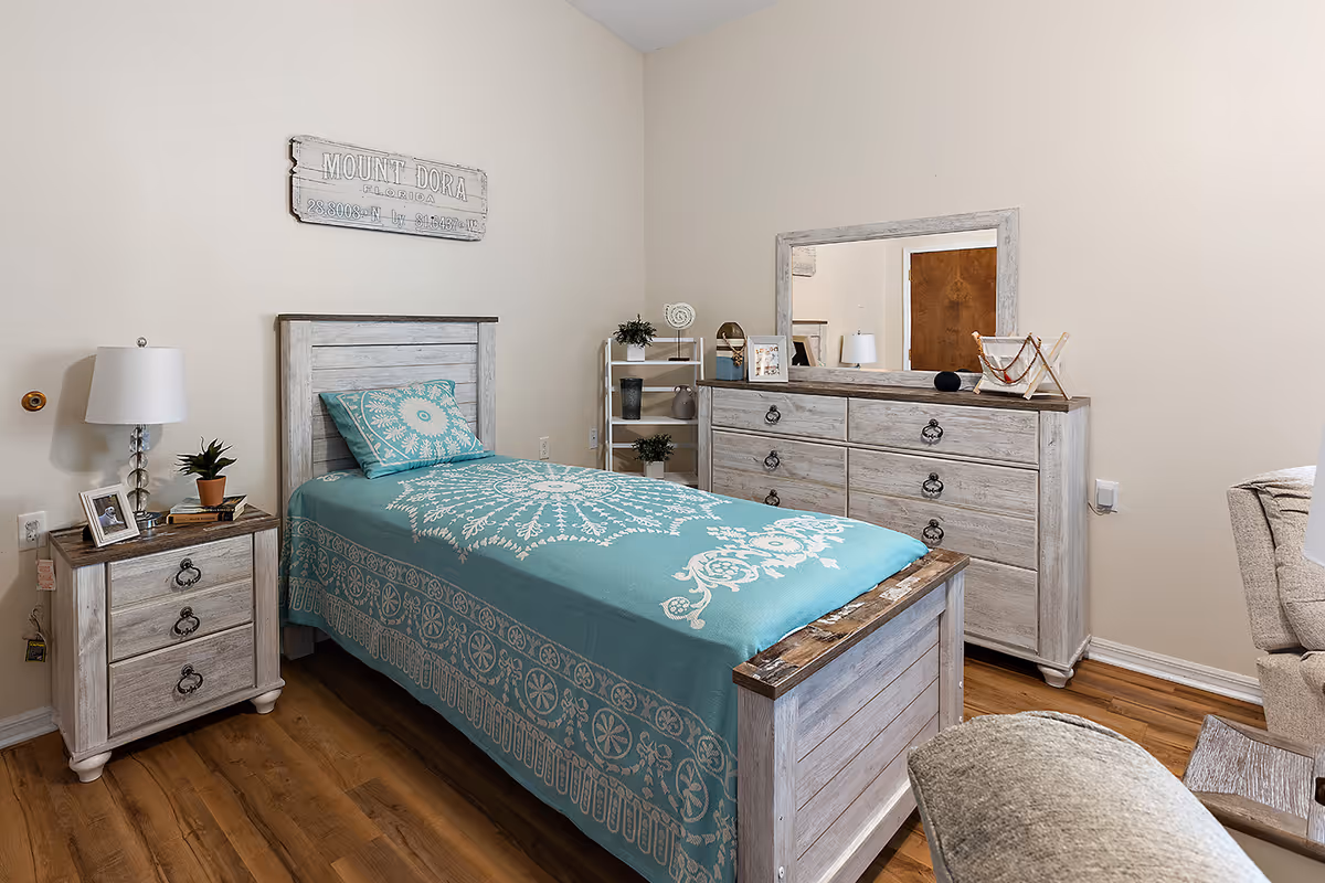 A furnished bedroom with a single bed covered in a teal patterned bedspread, whitewashed nightstand and dresser with mirror, and hardwood floor.