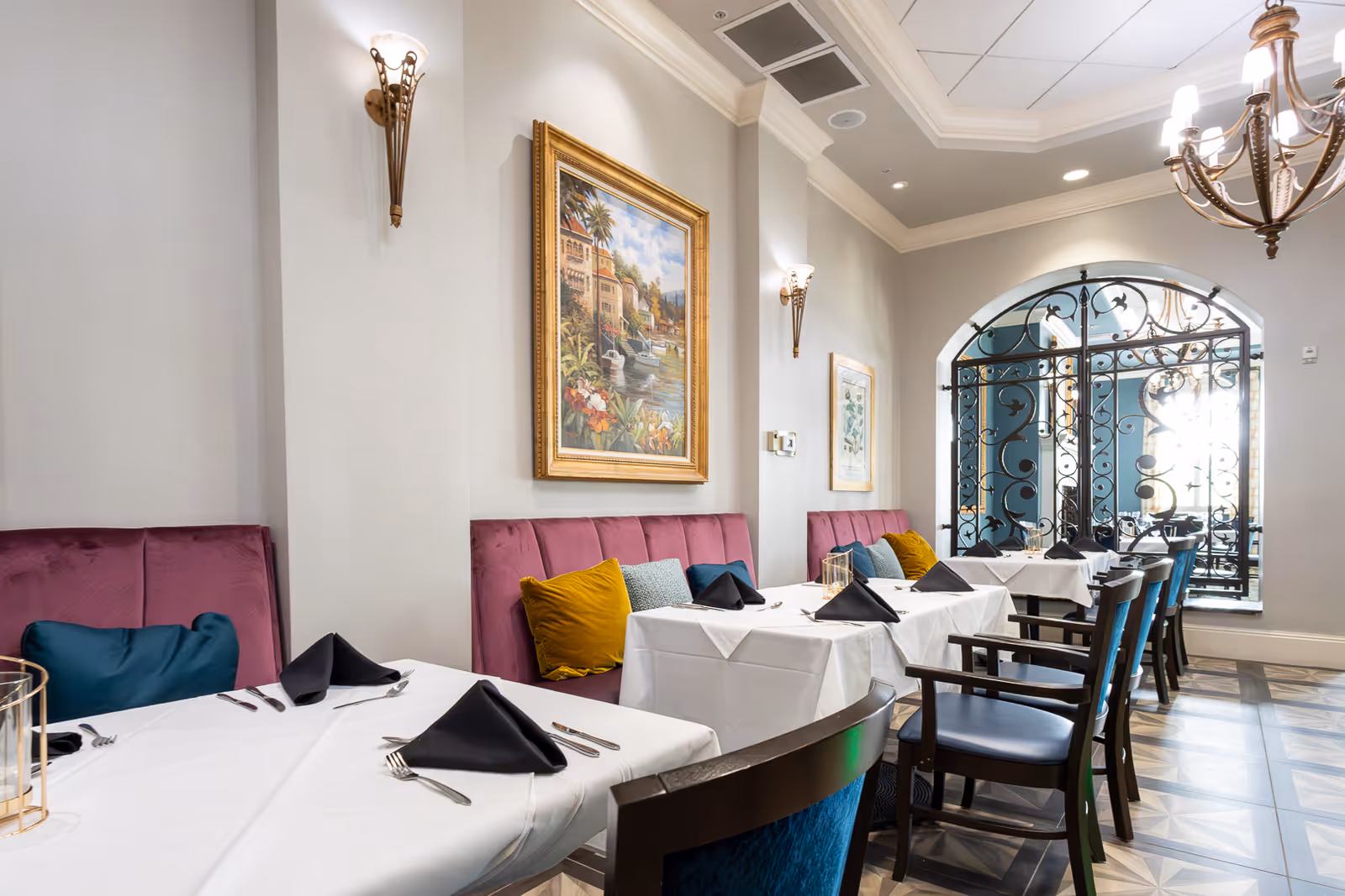Elegant dining room with white-clothed tables, upholstered banquette seating, framed artwork and decorative iron window details.