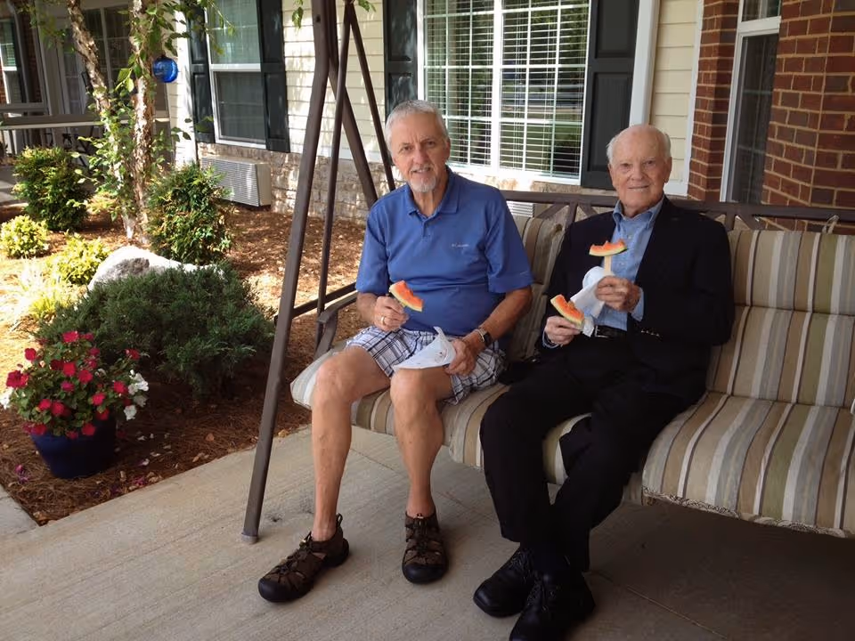 Two elderly men sitting on a striped cushioned outdoor swing on a patio, each holding a slice of watermelon. Behind them is a building with windows and some greenery including bushes and potted flowers.