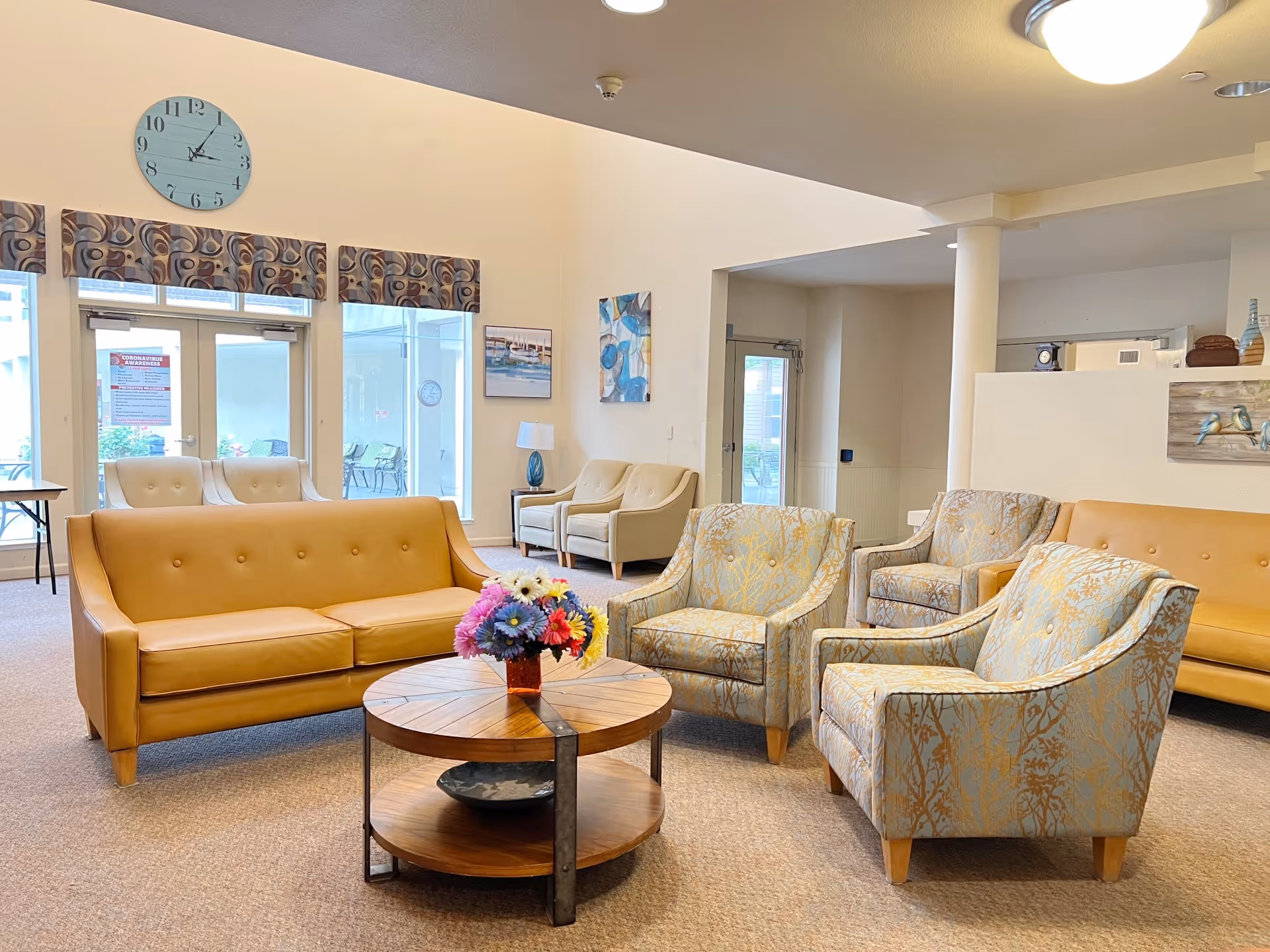 A bright and spacious living room area in a senior living facility with beige and patterned armchairs, two mustard yellow sofas, a round wooden coffee table with a vase of colorful flowers, large windows and glass doors letting in natural light, a blue wall clock, and decorative wall art.