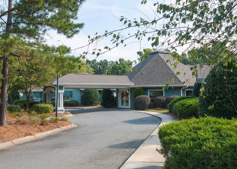 Exterior view of a senior living facility building with a driveway leading to a covered entrance. The building is surrounded by well-maintained bushes, trees, and landscaping under a clear sky.