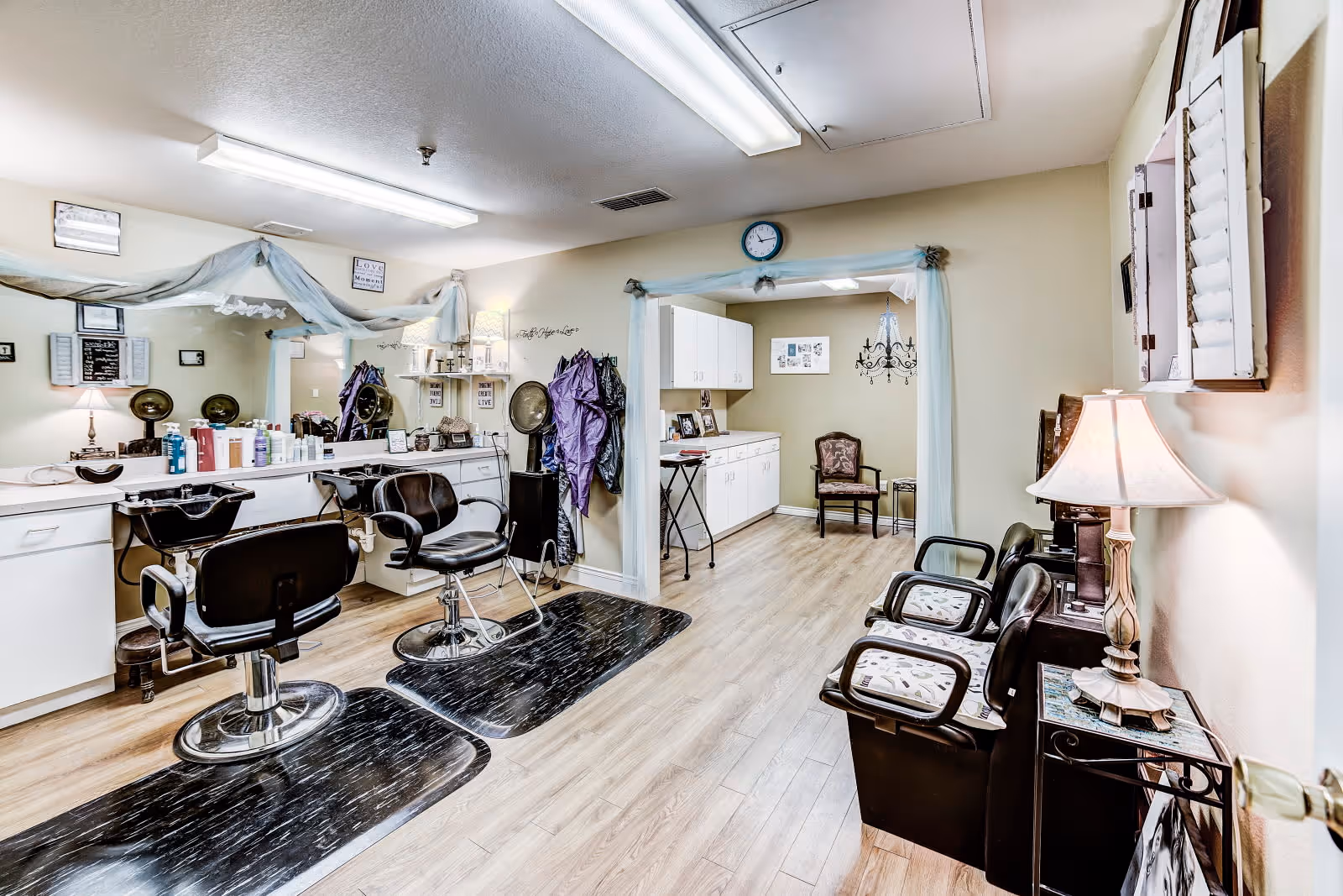 Interior view of a hair salon area within an assisted living facility. The room features two black salon chairs in front of a long counter with mirrors, hair care products, and hair dryers mounted on the wall. There are black mats on the wooden floor beneath the chairs. To the right, there is a waiting area with three black chairs with patterned cushions and a small table with a lamp. The walls are light-colored, and the room is well-lit with ceiling lights.