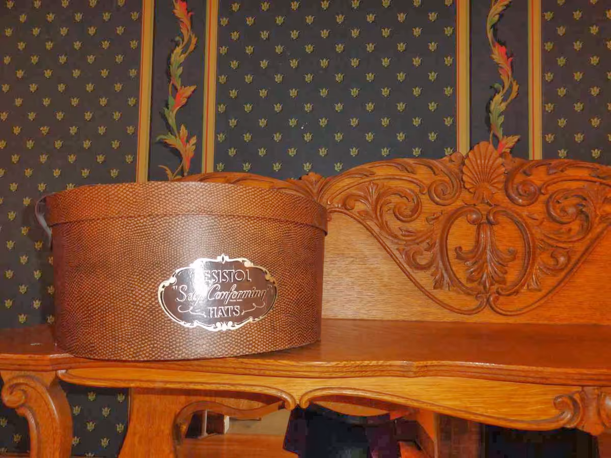 A close-up view of a wooden table with intricate carved details on the backrest. On the table, there is a brown textured hatbox labeled 'Bristol Self Conforming Hats'. The background features wallpaper with a dark base color and a floral pattern with vertical stripes.