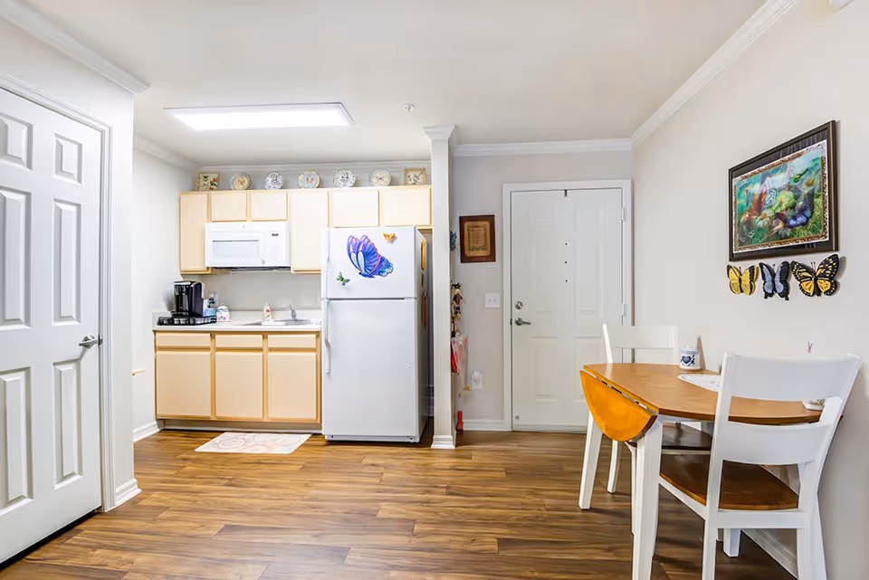 A small kitchen area with light wood cabinets, a white microwave above a countertop with a coffee maker, and a white refrigerator decorated with butterfly magnets. To the right is a wooden dining table with two white chairs, and a colorful butterfly-themed painting on the wall above it. The floor is wood, and there is a white door in the background.