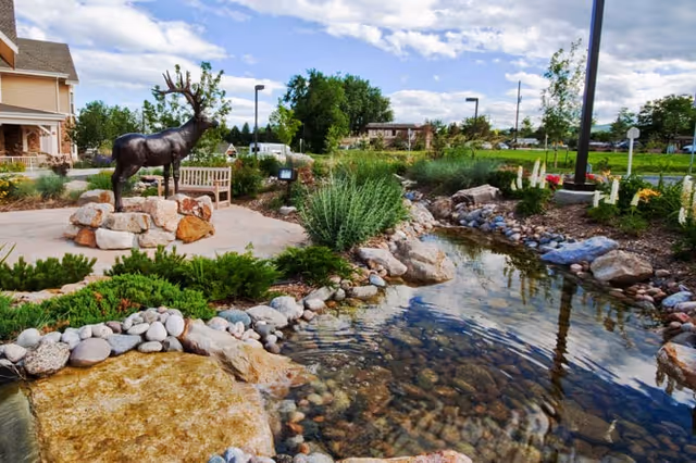 Landscaped outdoor courtyard featuring a clear rock-lined pond, shrubs and flowers, a bench, and a metal elk statue near a building under a partly cloudy sky.