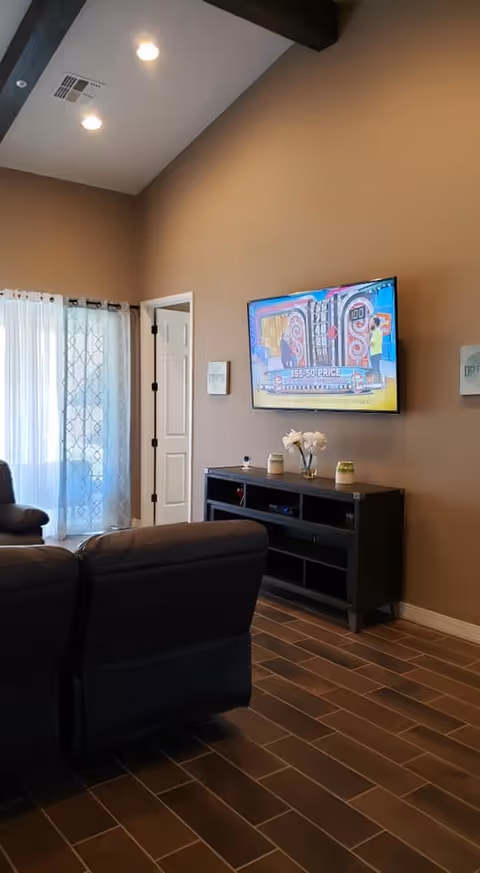 A cozy living room area with brown walls and wood-look tile flooring. There is a dark brown reclining sofa facing a wall-mounted flat-screen TV displaying a colorful game show. Below the TV is a black entertainment console with decorative items including a vase with white flowers and two small candles. A door and a window with sheer white curtains are visible in the background.