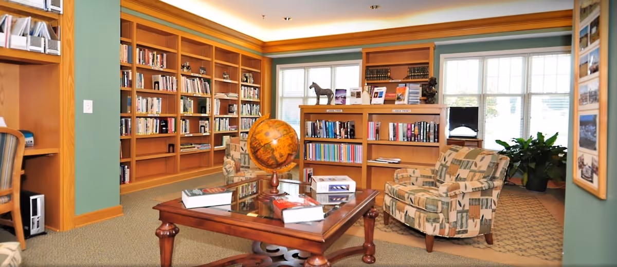 A cozy library room with wooden bookshelves filled with books, a wooden coffee table with a globe and several books on it, and two patterned armchairs. Large windows allow natural light to brighten the space, and there is a green plant near the window. The walls are painted green with wooden trim.