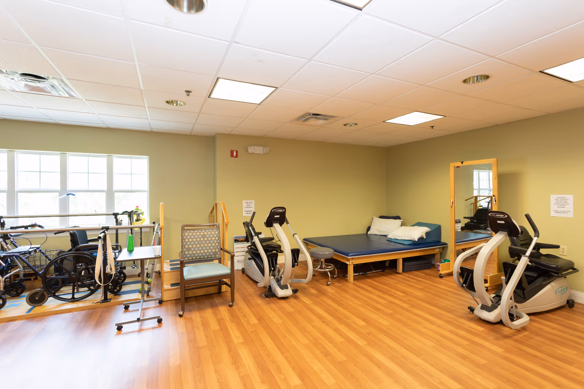A rehabilitation or therapy room with exercise equipment including two stationary bikes, a wheelchair, walkers, a padded therapy table with pillows, a chair, a small table, and a large mirror on the wall. The room has wooden flooring, beige walls, and a window letting in natural light.