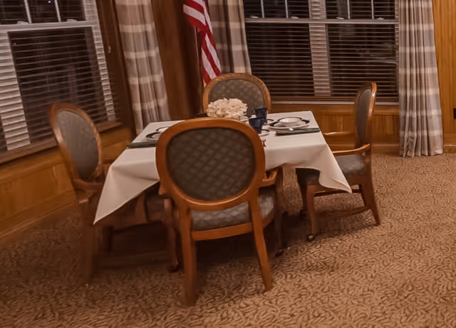 A dining area with a square table covered with a white tablecloth set for four people. Four wooden chairs with cushioned seats and backs surround the table. The room has carpeted flooring with a patterned design, large windows with blinds and plaid curtains, and an American flag in the corner.