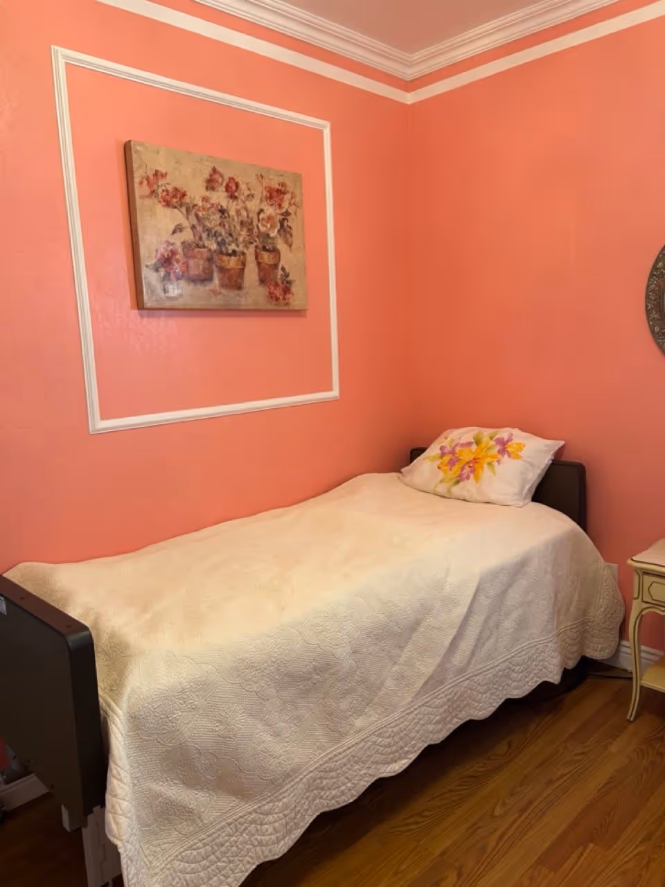 A small bedroom with coral pink walls and white trim. There is a single bed with a white quilted bedspread and a pillow with a floral design. Above the bed is a framed painting of flowers in pots. The floor is wooden, and a small white side table is partially visible next to the bed.