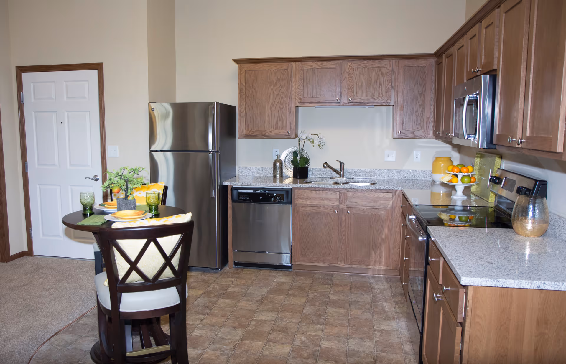 A modern kitchen with wooden cabinets, stainless steel refrigerator, dishwasher, microwave, and stove. There is a small round dining table with two chairs, set with green glasses, plates, and a small plant centerpiece. The floor is tiled, and the walls are painted light beige.
