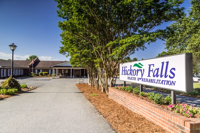 Exterior view of Hickory Falls Health and Rehabilitation facility with a large sign displaying the facility name in front. The building is single-story with a covered entrance, surrounded by trees, shrubs, and a clear blue sky.