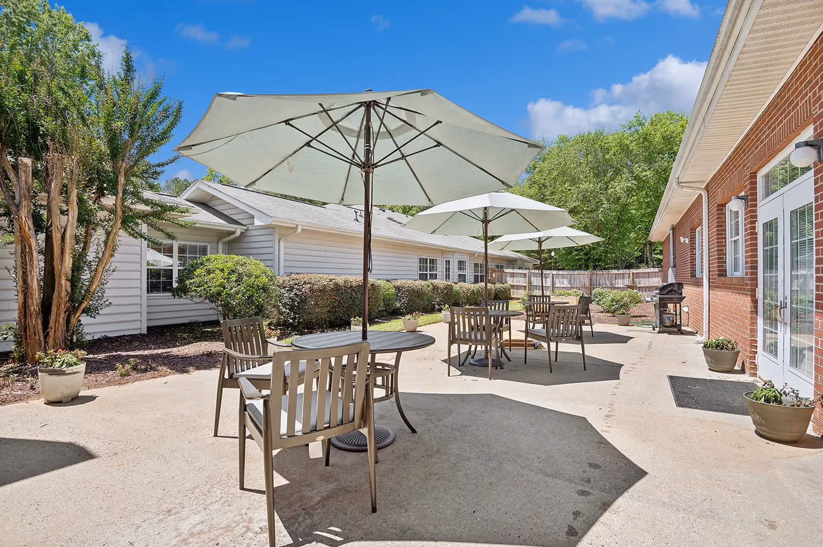 Outdoor patio area at Summit Place of Kings Mountain featuring several round tables with umbrellas and chairs. The patio is surrounded by greenery, including bushes and trees, and adjacent to a brick building with glass doors and potted plants.
