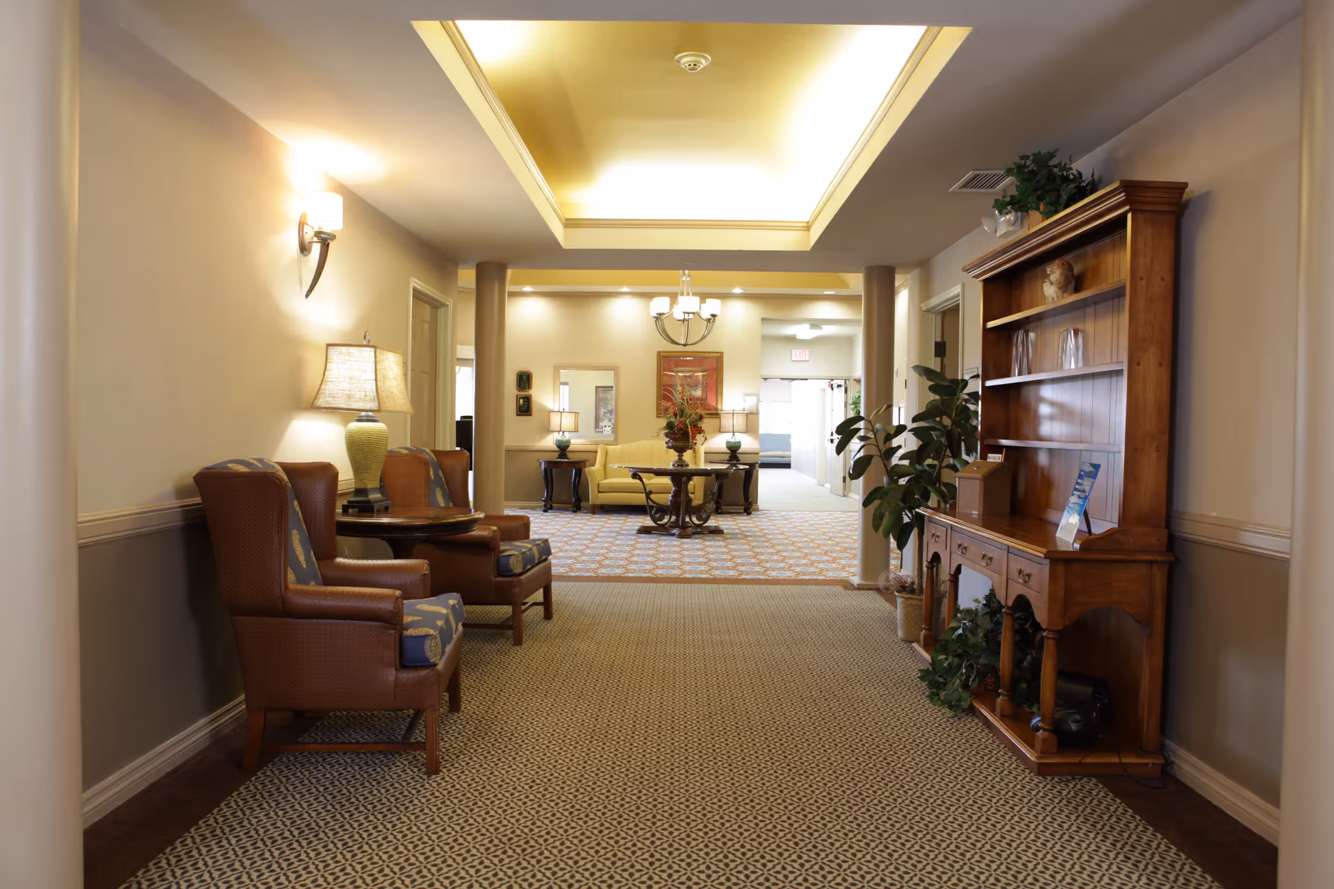 A well-lit senior living facility interior hallway with patterned carpet, two leather armchairs with a small round table and lamp on the left, and a wooden hutch with plants and decorative items on the right. In the background, there is a seating area with a yellow sofa, side tables with lamps, a round table with a flower arrangement, and a chandelier overhead.