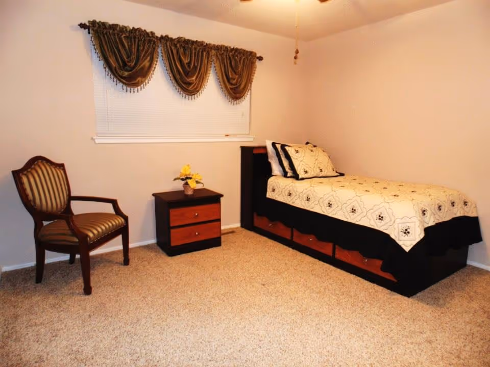 A simple bedroom with a single bed covered with a patterned beige and black bedspread, a wooden nightstand with two drawers and a small flower vase on top, a wooden chair with striped upholstery, and a window with brown draped curtains.