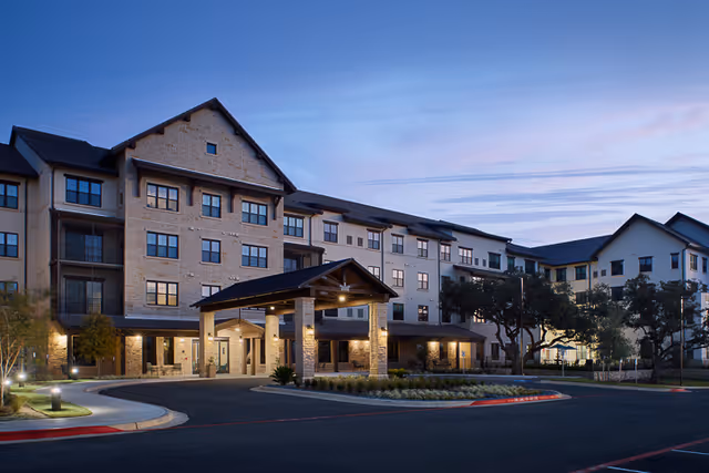 Exterior view of a large multi-story senior living facility building at dusk with illuminated entrance canopy, landscaped grounds, and a curved driveway.
