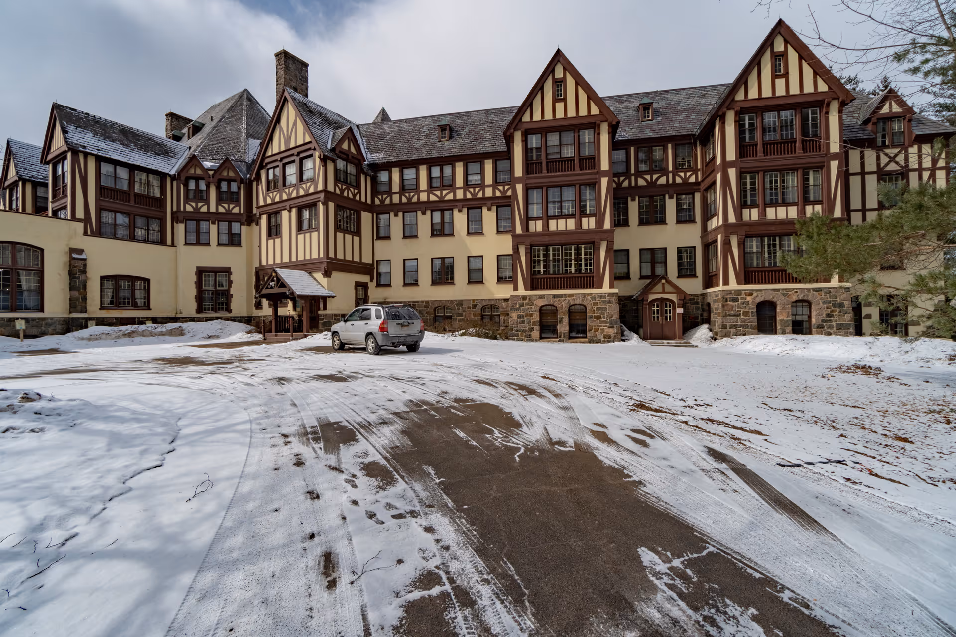 Exterior view of a large, Tudor-style building with multiple gables and windows, surrounded by snow-covered ground and a partially cleared driveway with a parked silver SUV.