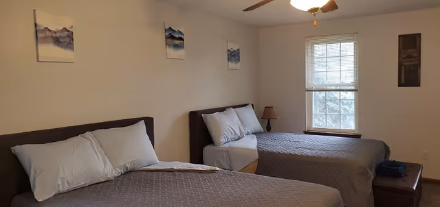 A bedroom with two neatly made beds with gray bedspreads, a window, ceiling fan, small bedside lamp, and wall artwork.