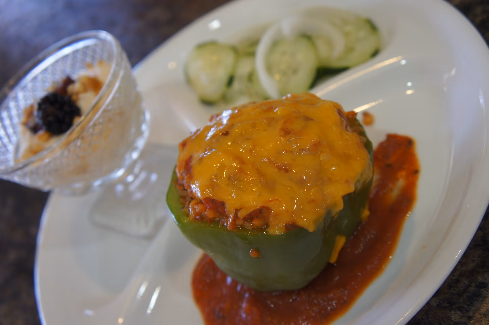 A close-up of a stuffed green bell pepper topped with melted cheddar cheese, served on a white plate with tomato sauce underneath. The plate also contains sliced cucumbers and onions, and a small glass bowl with a dessert topped with a blackberry and nuts.