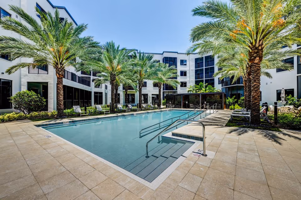 Courtyard swimming pool lined with palm trees and lounge chairs in the center of a multi-story senior living building.