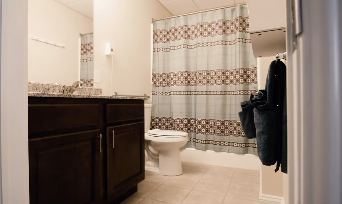 A bathroom with a granite countertop vanity with dark wood cabinets, a toilet, and a bathtub with a patterned shower curtain featuring blue and brown circular designs. There are dark towels hanging on a rack on the right wall and beige tiled flooring.
